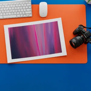 An unframed abstract fine art print is placed diagonally on an orange desk mat on top of a blue desk. A white keyboard, computer mouse, and a black digital camera are visible around the print