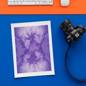 An unframed abstract fine art print is placed diagonally on an orange desk mat on top of a blue desk. A white keyboard, computer mouse, and a black digital camera are visible around the print.