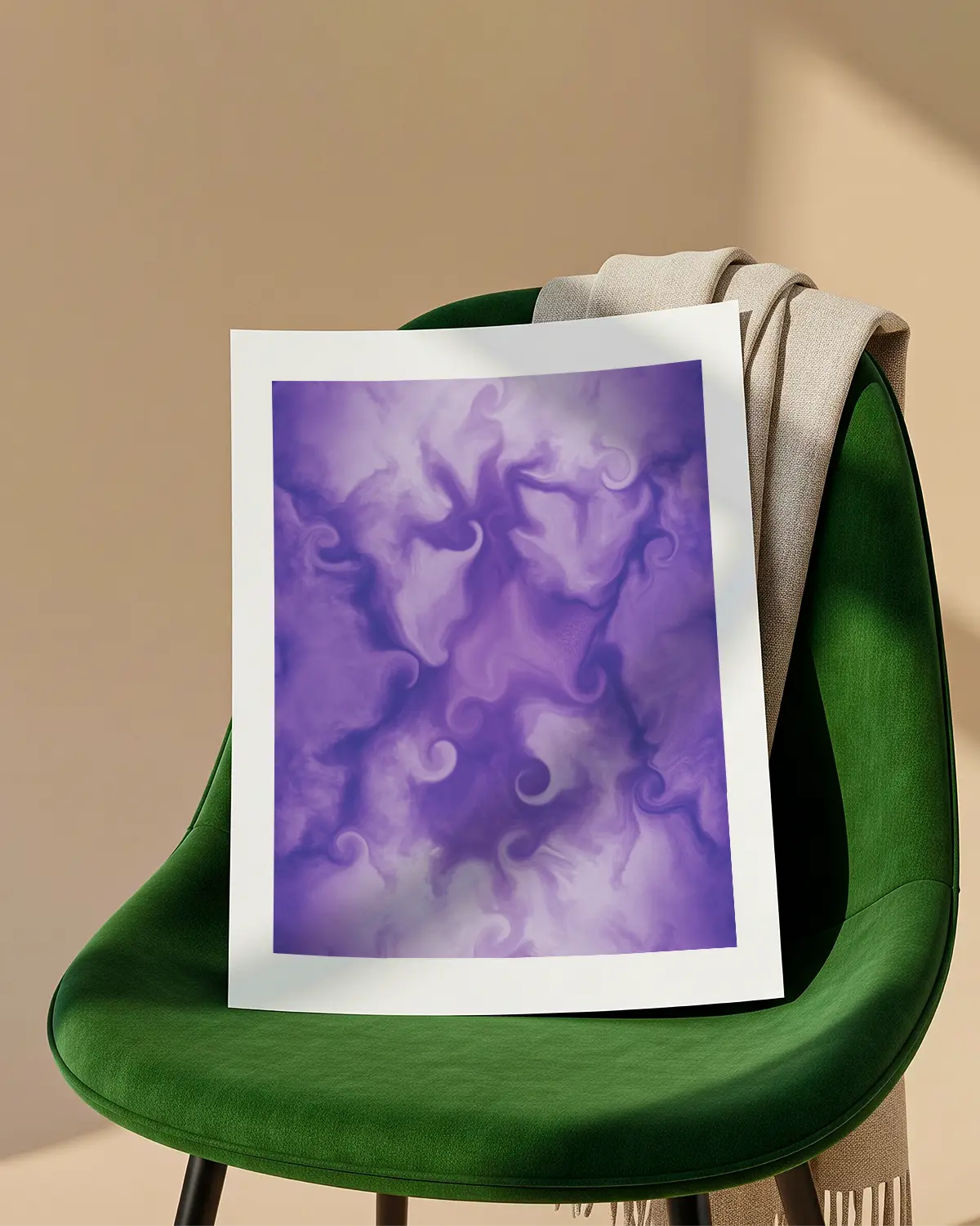 An unframed abstract fine art print with a clean white border is propped up on the seat of a green velvet chair. A beige blanket is draped over the back of the chair against a tan wall.