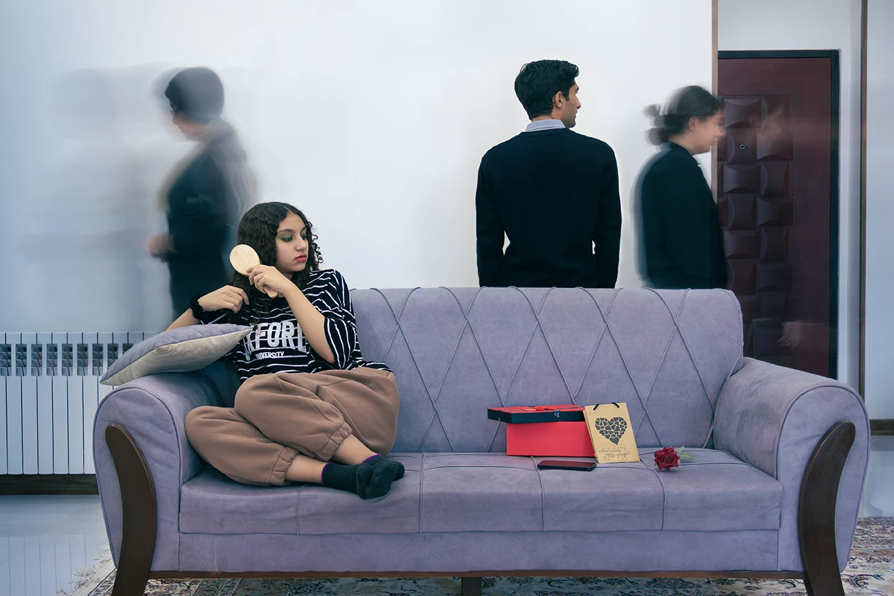 A woman sits on a purple couch, brushing her hair. Beside her are a small red gift box, a flower, and a mobile. Blurred figures walk past, creating a sense of time passing. A man stands behind the couch.