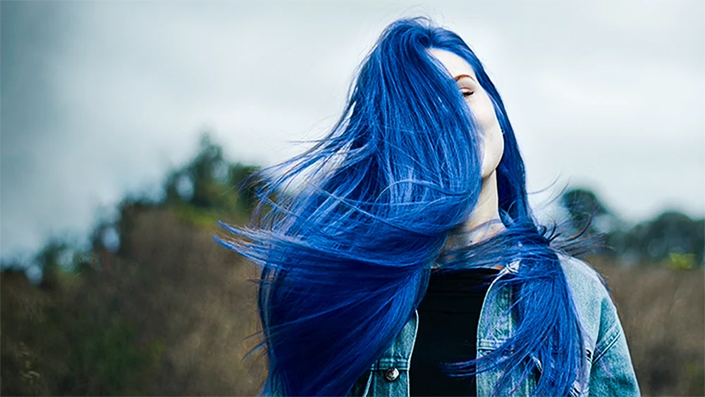 A girl with vibrant blue hair covers their face, wearing a denim jacket. The windy outdoor background is blurred